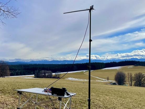 Zug der Alpen aus einer Hügel fotographiert, davor Funkgerät auf einem Tisch und kleiner Mast mit Yagi-Uda Antenne.
Die kleine Punkten im Himmel sind Heissluftballons.
